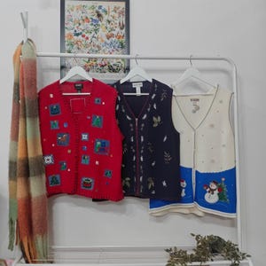 May include: Three decorative vests hang on a white rack. A red vest with square patches of Christmas designs, a navy blue vest with leaf embroidery, and a cream-coloured vest with a snowman and snowflake design. A colourful scarf hangs on the left.