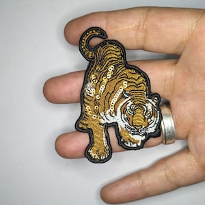 Peut inclure: Un écusson thermocollant brodé de couleur or et noir représentant un tigre avec des accents blancs. Le tigre est de face, la bouche fermée et les yeux ouverts. L'écusson est fait de paillettes et mesure environ 7,5 cm de haut.