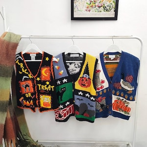Op de afbeelding: Drie mouwloze gebreide vesten hangend aan witte kledinghangers. Het linker vest is zwart met oranje randen en Halloween-motieven. Het middelste vest is veelkleurig met een pompoen- en kattenontwerp. Het rechter vest is blauw met een oogstthema met pompoenen.