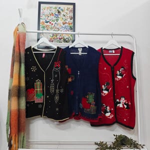May include: Three festive sweaters hang on a white rail. The sweaters are black, navy blue, and red. The black sweater features gift and ornament designs. The navy blue sweater has a zip and patchwork. The red sweater has penguin designs.