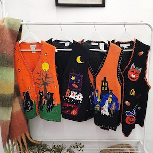May include: Four sleeveless knit vests hanging on white hangers. The first is orange with black cats and a tree. The second is black with a moon, stars, and the word BOO. The third is orange with a house and ghosts. The fourth is black with pumpkins.