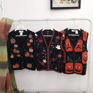May include: Three black knit V-neck sweater vests hanging on white hangers. The left vest features embroidered pumpkins. The middle vest displays a grid pattern with ghosts, pumpkins, and stars. The right vest shows large orange jack-o'-lantern faces.
