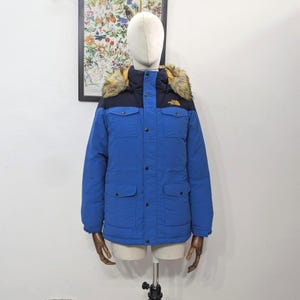 May include: A blue winter jacket with a black upper section and a faux fur-lined hood. The jacket has multiple pockets and a button-up front. The North Face logo is visible on the chest. The jacket is displayed on a mannequin.