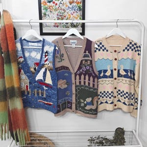 May include: Three knit vests and cardigans hanging on a white rack. The left vest features a blue nautical scene with a lighthouse and sailboat. The middle vest is tan with a garden and squirrel motif. The right cardigan is cream with a blue duck pattern.