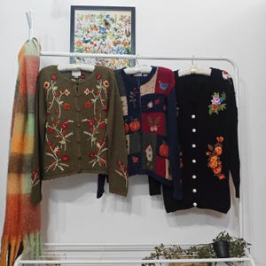 May include: Three embroidered cardigans hang on a white rack. The olive green cardigan has red and orange floral embroidery. The second cardigan is patchwork with fall-themed designs. The third is black with floral embroidery.