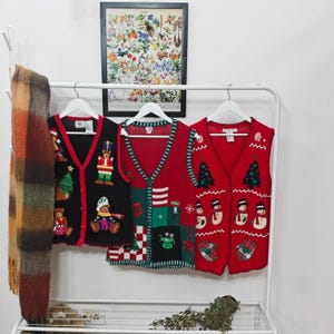 May include: Three Christmas-themed vests hang on a white rack. The vests are red and black, with festive designs including snowmen, Christmas trees, and teddy bears. A framed botanical print hangs above the rack.