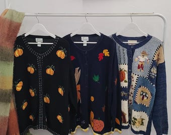 May include: Three long-sleeve button-down cardigans hanging on white hangers. The left is black with embroidered orange pumpkins. The center is navy blue with embroidered autumn leaves and pumpkins. The right features a patchwork design with a scarecrow.