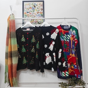 May include: Three Christmas sweaters hang on a white rack. One is black with green Christmas trees, another is black with white polar bears, and the third is red with gift boxes. A colorful scarf hangs on the left.