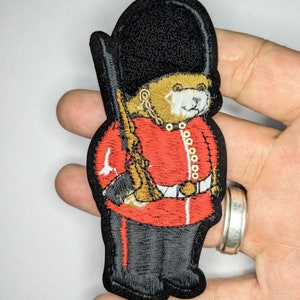 Peut inclure: Patch brodé représentant un ours en peluche habillé en garde britannique. L'ours porte une tunique rouge, un pantalon noir et un chapeau de peau d'ours noir. L'ours tient un fusil.