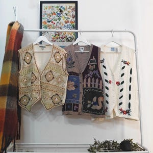 Peut inclure: Trois gilets vintage sont suspendus à un portant blanc. Les gilets comprennent un modèle en crochet, un modèle patchwork avec un écureuil et un gilet en tricot torsadé avec des broderies florales. Une écharpe colorée est suspendue à gauche.