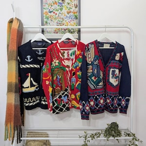 Puede incluir: Tres cárdigans vintage cuelgan de un perchero blanco. Uno es azul marino con un diseño de velero y ancla. El segundo es rojo con un patrón colorido y detallado. El tercero es azul marino con un escudo y patrones geométricos.