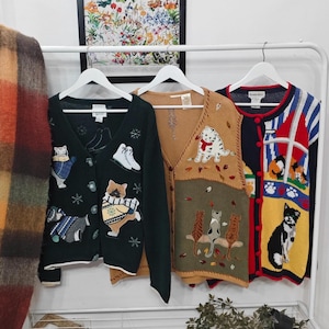 Könnte beinhalten: Drei Pullover mit Katzenmotiven hängen an einem weißen Gestell. Eine dunkelgrüne Strickjacke mit Katzen- und Schneeflockenapplikationen, eine beige Weste mit einer Katze mit Schal und ein blau-rot-gelber Pullover mit Katzenmotiven.