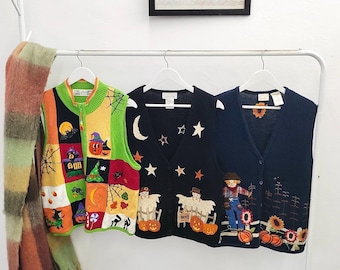 Puede incluir: Tres chalecos de punto sin mangas colgados en un perchero blanco. El chaleco izquierdo presenta un diseño de patchwork colorido con motivos de Halloween como calabazas, brujas y telarañas. El chaleco central es negro con patrones de luna y estrellas. El chaleco derecho es azul marino con un diseño de espantapájaros y girasoles.