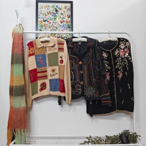 Pode incluir: Exibição de roupas vintage em um cabide branco. Os itens incluem um suéter bege com zíper e patchwork, uma jaqueta preta bordada e um cardigã preto com bordados florais. Um cachecol colorido está pendurado à esquerda.