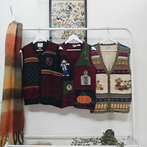 May include: Three vintage knit vests with distinctive patterns and designs are displayed on a white rack. The vests feature burgundy, green, and cream colours, with embroidered details such as a scarecrow, pumpkin, and geometric shapes. A patterned scarf hangs on the left.