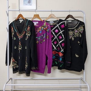 May include: Four long-sleeved sweaters hang on a white clothes rack. The sweaters are black and purple, adorned with colourful sequin and bead embellishments. One black sweater has a V-neck, while another features a diamond pattern. The sweaters are vintage and have a decorative style.