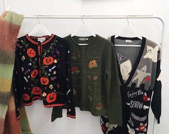 May include: Three long-sleeved cardigans hanging on white hangers. The left is black with orange pumpkin and candy embroidery. The center is olive green with floral and leaf embroidery. The right is black and gray with ghost graphics and the text Enjoy the SHOW.