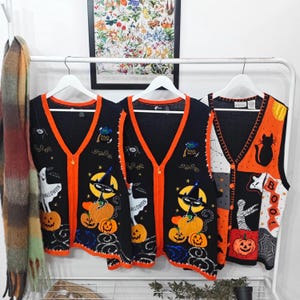 May include: Three sleeveless V-neck sweater vests hanging on white hangers. Two black vests feature orange trim, a cat in a witch hat on a pumpkin, and a ghost. The third vest is color-blocked in black, orange, and white with a cat and pumpkin design.