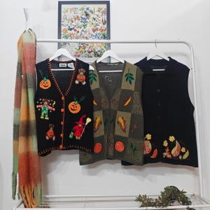 May include: Three vintage vests hang on a white rack. A black vest with Halloween embroidery, a green patchwork vest, and a black vest with floral embroidery. A colourful scarf hangs on the left.