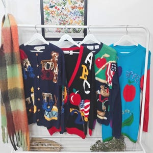 Könnte beinhalten: Eine Sammlung von Vintage-Sweatern und einem Schal, die auf einem weißen Kleiderständer hängen. Die Pullover zeigen farbenfrohe Designs, darunter Äpfel, die Buchstaben ABC und Teddybären. Der Schal ist eine Mischung aus Orange, Braun und Grün.