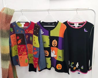 May include: Three long-sleeve sweaters hanging on white hangers. The left sweater features a multi-colored patchwork design with autumn leaves and pumpkins. The middle sweater has a black, purple, and green block pattern with pumpkins and ghosts. The right sweater is black with small embroidered Halloween motifs.