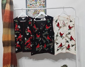 May include: Three knitted vests with cardinal bird designs. One black vest and one cream vest are button-up, and the other black vest is a pullover. The vests feature red cardinals, green leaves, and white floral accents.