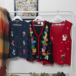 May include: Three Christmas-themed vests on white hangers. The vests are navy blue, black, and red. The navy vest has Christmas stocking and holly embroidery. The black vest has gift box and tree embroidery. The red vest has star and Santa embroidery.