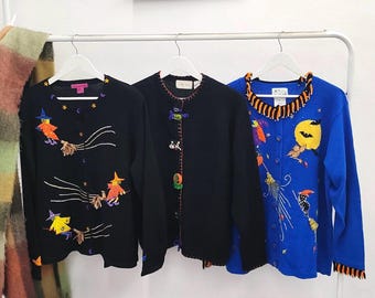 Puede incluir: Tres cárdigans de manga larga con botones en perchas blancas. El de la izquierda es negro con brujas bordadas en escobas. El central es negro con pequeños apliques de Halloween. El de la derecha es azul con luna, murciélago y bruja.