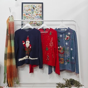 May include: Three festive Christmas sweaters on a white rail. The sweaters are navy, red, and blue. The navy sweater features a Santa Claus design. The red sweater has a Christmas tree design. The blue sweater has a sleigh design.