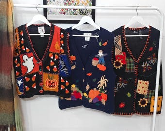 May include: Three sleeveless knit vests hanging on white hangers. The left vest is black with ghost, pumpkin, and candy corn motifs. The center navy blue vest features a scarecrow and harvest vegetables. The right black vest has sunflower and plaid patches.