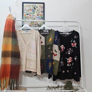 May include: A clothes rail holds a beige cardigan, a patchwork zip-up sweater, and a black floral top. A fringed scarf in orange, brown, and green hangs on the left. A framed botanical print hangs above the rail.