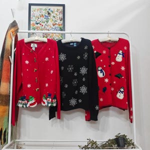 May include: Three Christmas sweaters hang on a white rack. A red cardigan with snowman and snowflake designs, a black crewneck sweater with snowflakes, and a red cardigan with snowmen and snowflakes.