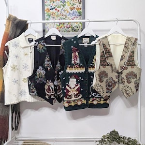 May include: Four festive vests hang on a white rack. The vests feature holiday-themed designs, including snowflakes, Christmas trees, and Santa Claus. Colors include cream, black, green, and tan. The vests are made of various materials and textures.
