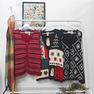 May include: Three sleeveless sweaters hang on a white rail. The sweaters are red, black, and a patchwork design with Christmas motifs. A colourful scarf hangs on the left, and a framed botanical print is in the background.