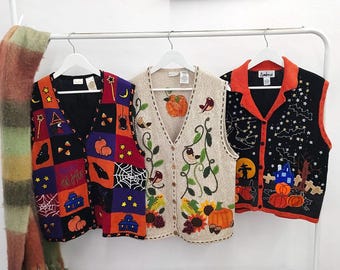 May include: Three sleeveless knit vests hanging on a white rack. The left vest features a colorful patchwork pattern with Halloween motifs. The center vest is beige with embroidered autumn foliage and birds. The right vest is black with orange trim.