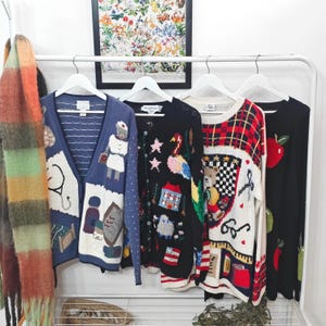 Könnte beinhalten: Eine Sammlung von Vintage-Sweatern auf einem Kleiderständer. Die Pullover weisen verschiedene Designs auf, darunter eine blaue Strickjacke mit medizinischer Stickerei, ein schwarzer Pullover mit einem Vogel und einem Haus sowie ein rot-weiß karierter Pullover.
