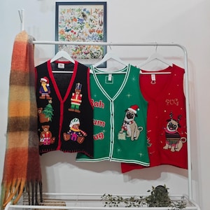 May include: Three Christmas-themed vests hang on a white rack. The vests are black, green, and red, each with unique holiday designs. A scarf hangs on the left. A framed botanical print hangs in the background.