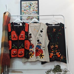 May include: Three autumn-themed vests hang on a white rack. One black vest features orange jack-o'-lanterns. A beige vest has scarecrows and pumpkins. The third black vest has autumn leaves and pumpkins. A plaid scarf hangs on the left.