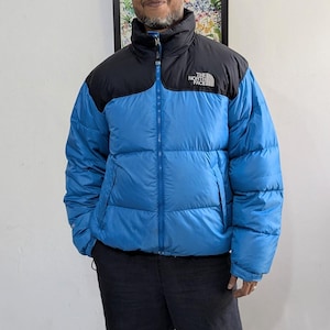 May include: A blue and black puffer jacket with a high collar and a zip fastening. The North Face logo is visible on the upper chest. The jacket has a quilted design and appears well-padded for warmth. The jacket is worn by a person.