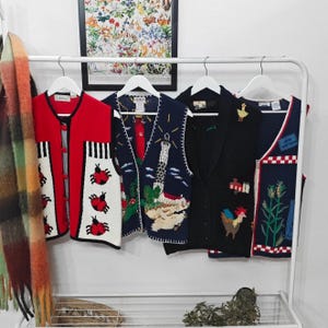 Puede incluir: Una colección de chalecos de punto vintage en varios colores y diseños. Los chalecos incluyen uno rojo con mariquitas, uno azul marino con un faro, uno negro con un gallo y uno azul marino con mazorcas de maíz. Están colgados en un estante blanco.