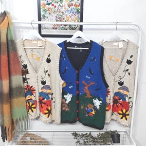 May include: Three sleeveless knit vests hanging on a white rack. Two beige vests feature scarecrow, pumpkin, and ghost motifs. The centre blue vest displays a Halloween scene with a bear, ghosts, a tree, and the word Boo printed on the fabric.