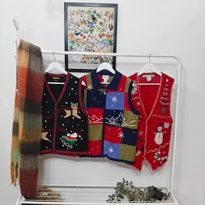 May include: Three festive Christmas sweaters hang on a white rack. One is black with reindeer and Santa, another is red, blue, and green with a winter scene, and the third is red with a snowman and Christmas tree.