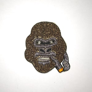 May include: Embroidered patch of a gorilla's face with a cigarette in its mouth. The gorilla is brown and the cigarette is orange and white.