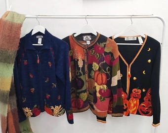 May include: Three knit cardigans hanging on white hangers. The left is navy blue with embroidered autumn motifs. The center features a multi-colored harvest pattern with pumpkins and wheat. The right is black with orange trim and jack-o'-lantern designs.