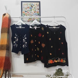 May include: Three sleeveless cardigans hang on a white rail. The first is navy blue with white floral embroidery. The second is black with gold buttons and embroidered autumn leaves. The third is black with embroidered pumpkins and leaves.