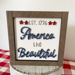 May include: A framed wood sign with the text "EST. 1776" and "America the Beautiful" in blue lettering. The sign has a white background and is decorated with three red stars.