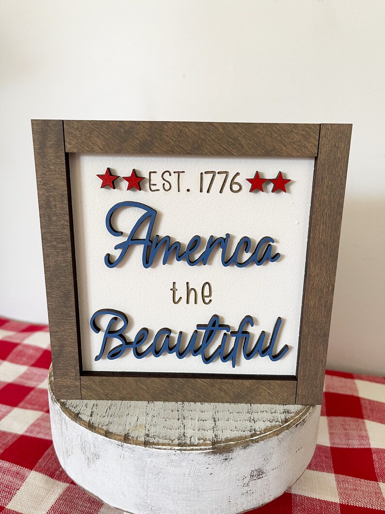 May include: A framed wood sign with the text "EST. 1776" and "America the Beautiful" in blue lettering. The sign has a white background and is decorated with three red stars.