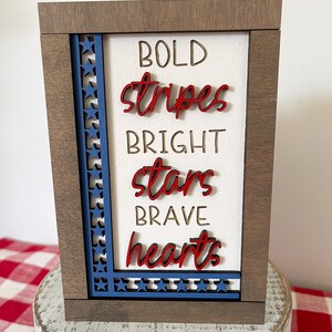 May include: A wooden sign with a white background and a blue and red star border. The sign says "BOLD stripes BRIGHT stars BRAVE hearts".