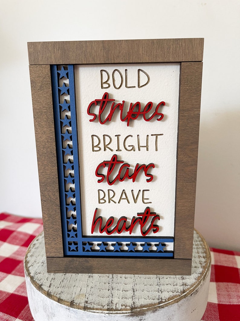 May include: A wooden sign with a white background and a blue and red star border. The sign says "BOLD stripes BRIGHT stars BRAVE hearts".