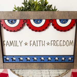 May include: A wooden framed sign with a white background and the words "FAMILY * FAITH * FREEDOM" in a cutout font. The sign is decorated with red, white, and blue stars and banners.